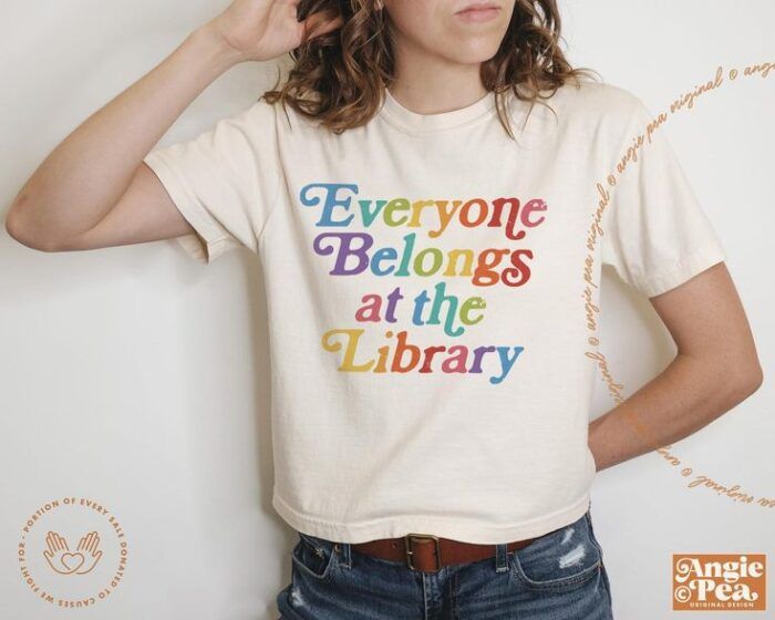 White, slim-fit crop top with rainbow font that reads "everyone belongs at the library."