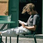 fair-skinned man resting his feet on a cafe chair outside and reading a book