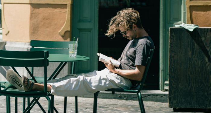 fair-skinned man resting his feet on a cafe chair outside and reading a book