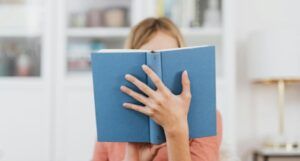 fair-skinned person holding a light blu book in front of their face