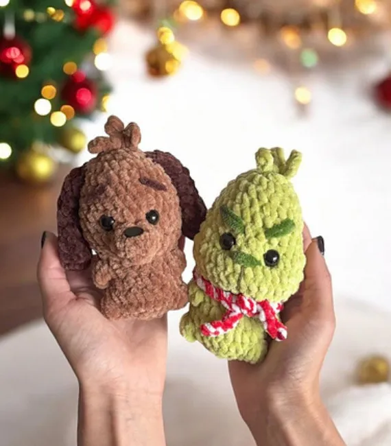 Two little crocheted amigurimi figurines designed to look like Max, a little brow dog with floppy ears, and the Grinch, a green, pointy headed creature with a red scarf, from How the Grinch Stole Christmas are being held out in front of a Christmas tree.