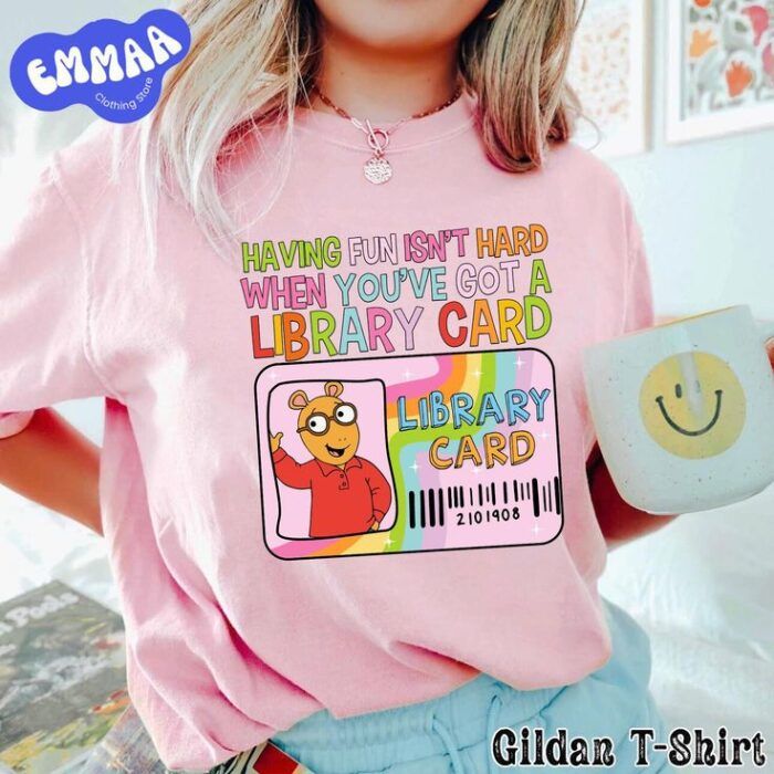 a pink t-shirt with an image of the cartoon character arthur on a library card and rainbow font that reads "having fun isn't hard when you've got a library card."