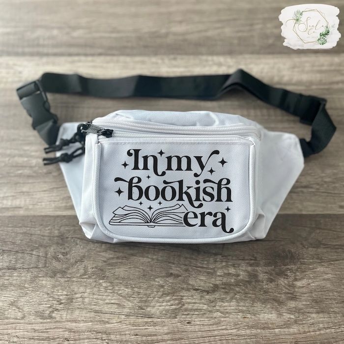 white fanny pack with a black strap. the fanny pack has black text that reads "in my bookish era" and a graphic of an open book in black ink