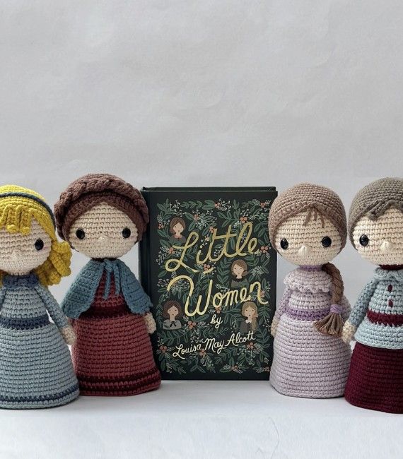 Four little crocheted aigurumi figures wearing historical dresses in the style of the American Civil War and designed to look like Amy, Meg, Beth, and Jo from Little Women. The figures are set around a beautiful green edition of the book, The Little Women.