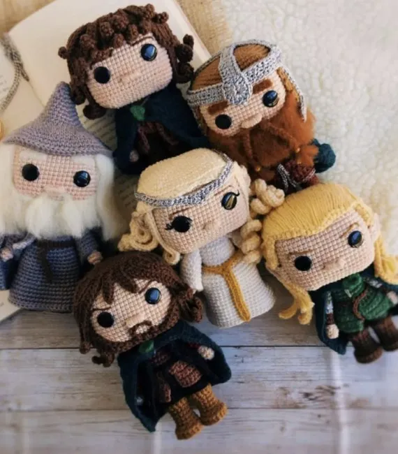 A whole collection of crocheted figures from The Lord of the Rings are grouped together against the open pages of a book. Their heads are slightly square and shaped like Funko Pops. There's Gandalf, Frodo, Gimli, Aragorn, Galadriel, and Legolas.