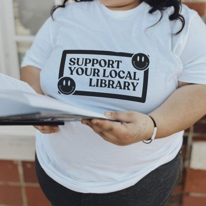 a white t-shirt with a simple, black, retro-inspired design including the words "support your local library" and smiley faces 