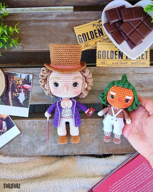 Crocheted amigurumi figurine of Willy Wonka with a purple jacket and brown top hat alongside a tiny orange oompa loompa being held out in a white woman's palm.