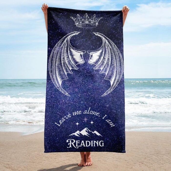 image of a person on a beach holding up a royal blue beach towel with images of wings, a crown, and mountains around the words "leave me alone, I am reading."