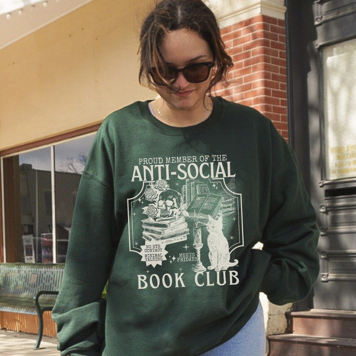 Image of a person in a dark green crewneck sweatshirt with a white illustration of a book stack and a cat with text "members only anti-social book club -- no eye contact, minimal talking, meets on fridays"
