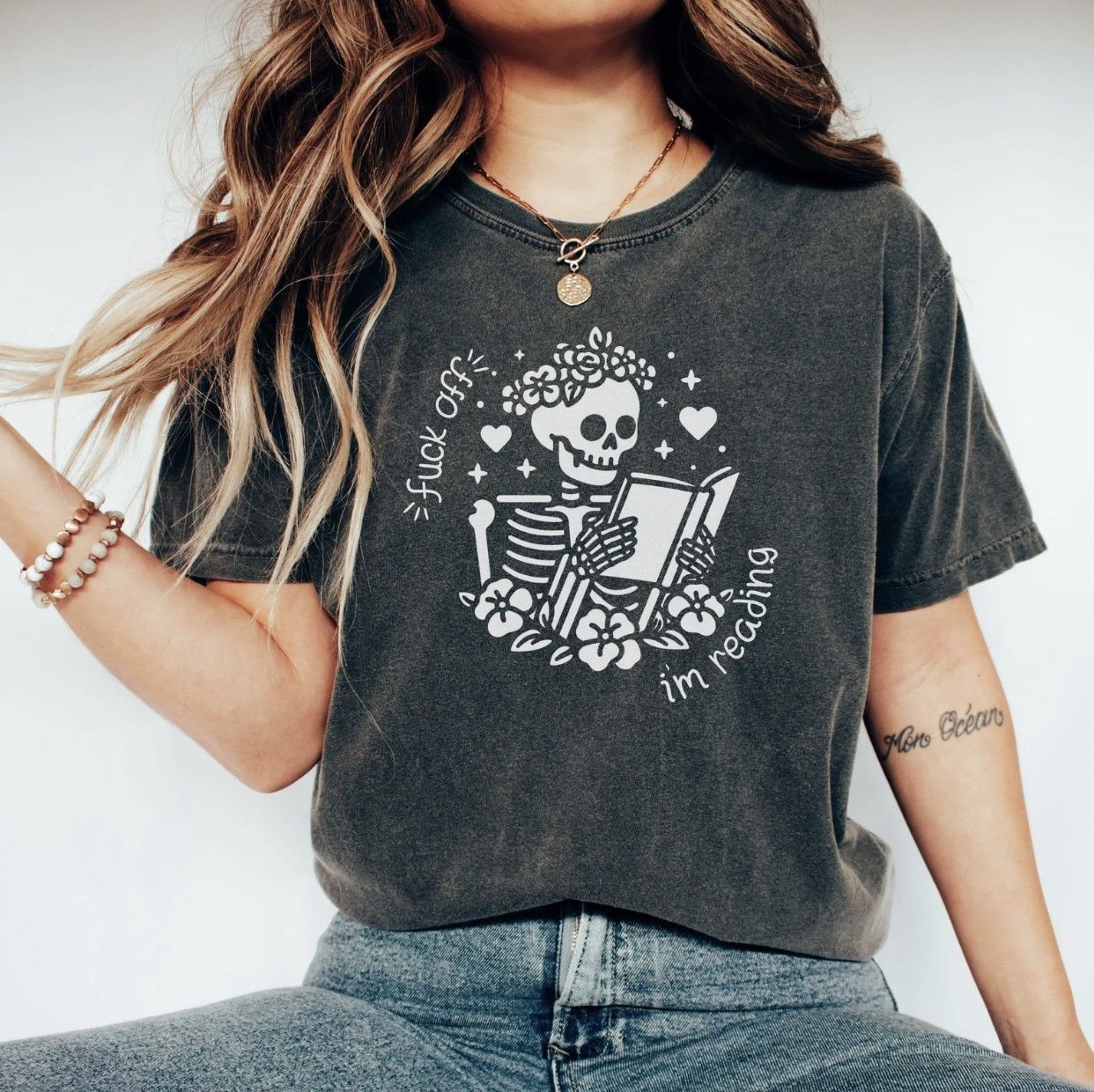 Image of someone in a dark gray t-shirt with an illustrated skeleton reading a book and the text "fuck off, I'm reading"