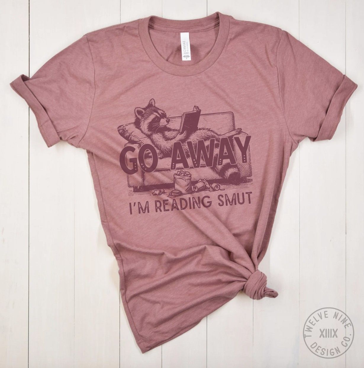 Image of a pink t-shirt with an illustration of a raccoon reading a book on a couch with the text "go away, I'm reading smut"