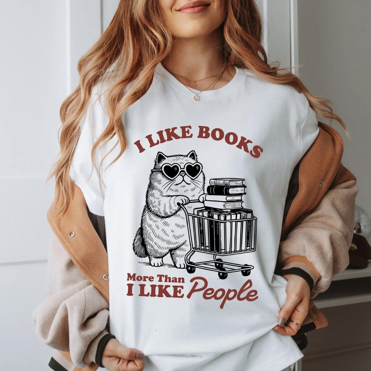 Image of a person in a white t-shirt with an illustration of a cat with heart-shaped sunglasses pushing a cart of books and the text "I like books more than I like people"