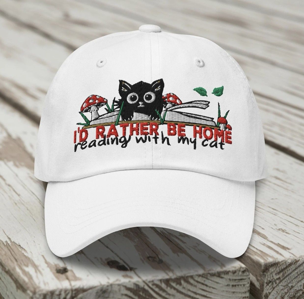 Image of a white baseball cap embroidered with an illustration of a cat and books and the words "I'd rather be home reading with my cat"