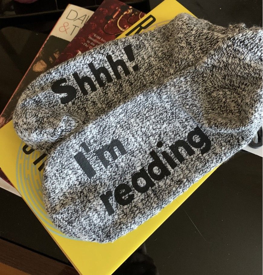 Image of a pair of gray knit socks with words ironed on the bottoms "Shhh! I'm reading"