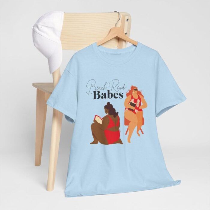 a shirt and a white baseball cap hanging on a chair. The shirt has two plus sized women, one Black, one white, in bathing suits, holding books. The words read "Beach Read Babes"