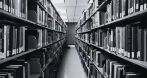 black and white image of a library aisle with shelves of books and brick wall at end