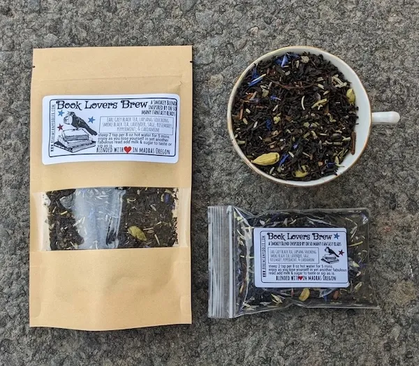 two bags of tea, one larger brown one, one small clear one, with labels for Book Lovers Brew. The bags are arranged next to a cup full of loose leaf black tea