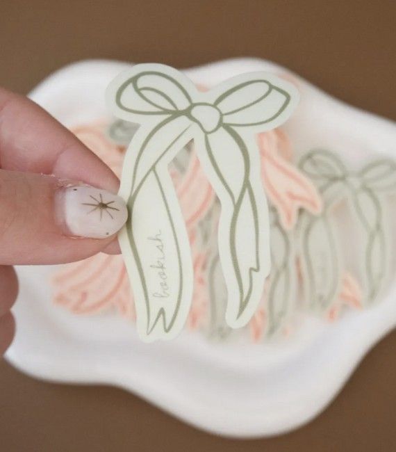 A white woman's fingers hold up a sticker of an illustrated green ribbon with the word "bookish" written on one side of the long ribbons