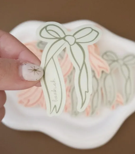 A white woman's fingers hold up a sticker of an illustrated green ribbon with the word "bookish" written on one side of the long ribbons