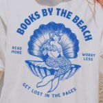 image of a white person with blonde hair in a ponytail, standing on a beach. They are wearing a white shirt with a blue design of a vintage mermaid in a sea shell and the words Books by the Beach