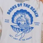image of a white person with blonde hair in a ponytail, standing on a beach. They are wearing a white shirt with a blue design of a vintage mermaid in a sea shell and the words Books by the Beach