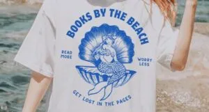 image of a white person with blonde hair in a ponytail, standing on a beach. They are wearing a white shirt with a blue design of a vintage mermaid in a sea shell and the words Books by the Beach
