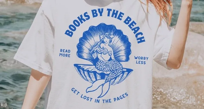 image of a white person with blonde hair in a ponytail, standing on a beach. They are wearing a white shirt with a blue design of a vintage mermaid in a sea shell and the words Books by the Beach