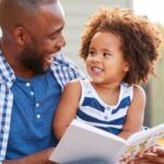 a brown-skinned father and child reading a book together and laughing