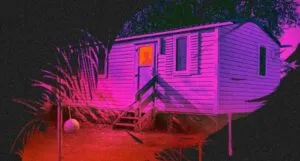 cropped book cover of Mapping the Interior by Stephen Graham Jones showing a person in the window of a modular home. the image is bathed in purple, pink, red, and black