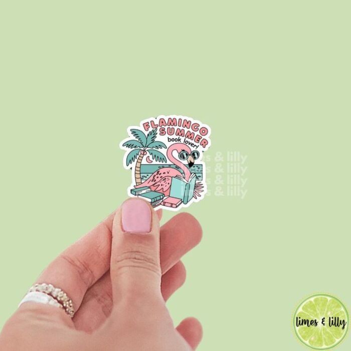a white hand wearing a silver ring holding a small sticker showing a flamingo wearing sunglasses and reading on a beach with the words "Flamingo Summer Book Lover"