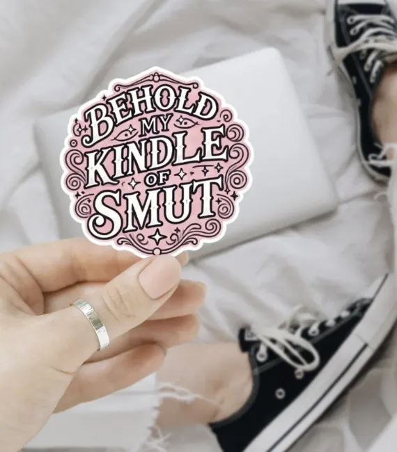A pink sticker with filagree doodles around the edges and white letters outlines in black that reads "Behold my kindle of smut" being held up between a woman's fingers
