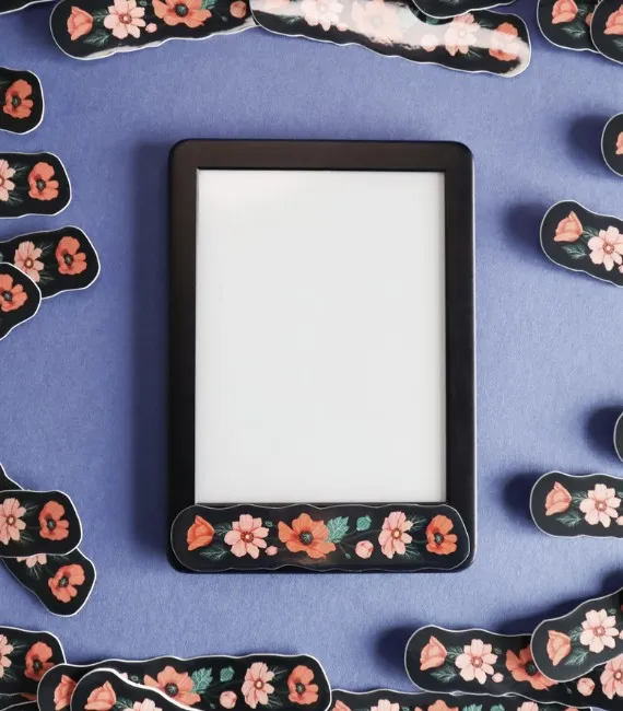 A kindle set against a purple background is surrounded by floral stickers. The sticker features red and pink flowers in a line against a black background.