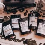 six black resealable pouches of loose leaf teas with white labels for Lord of the Rings themed tea. The blends are Gondor, Lothlorien, Misty Mountain, Rivendell, Rohan, and Shire, and the bags are on a table with a stack of Lord of the Rings books and assorted ceramics
