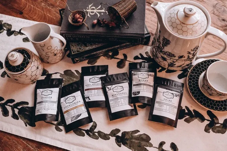 six black resealable pouches of loose leaf teas with white labels for Lord of the Rings themed tea. The blends are Gondor, Lothlorien, Misty Mountain, Rivendell, Rohan, and Shire, and the bags are on a table with a stack of Lord of the Rings books and assorted ceramics