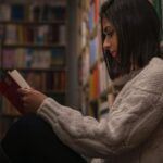 pale-skinned woman with a bob sitting in library reading between the stacks