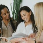 3 women, of various skin tones and ethnicities, stand together smiling and looking at a book