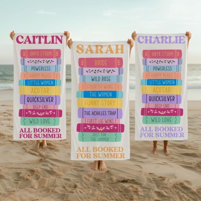 three people standing on a beach holding up white beach towels personalized with names and stacks of books in bright colors 