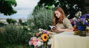 a person with pale skin and long red hair reading in a garden