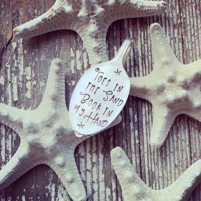 weathered wood and bleached starfish surround a vintage spoon with a folded handle and the words "toes in the sand, book in my hand" engraved on the spoon head