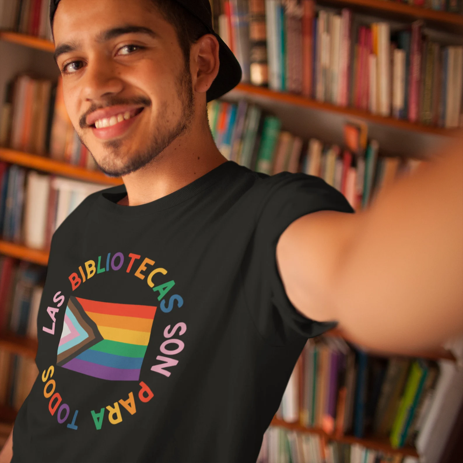 Image of a brown person wearing a black shirt that says "las bibliotecas son para todos" with a rainbow flag in the center. 