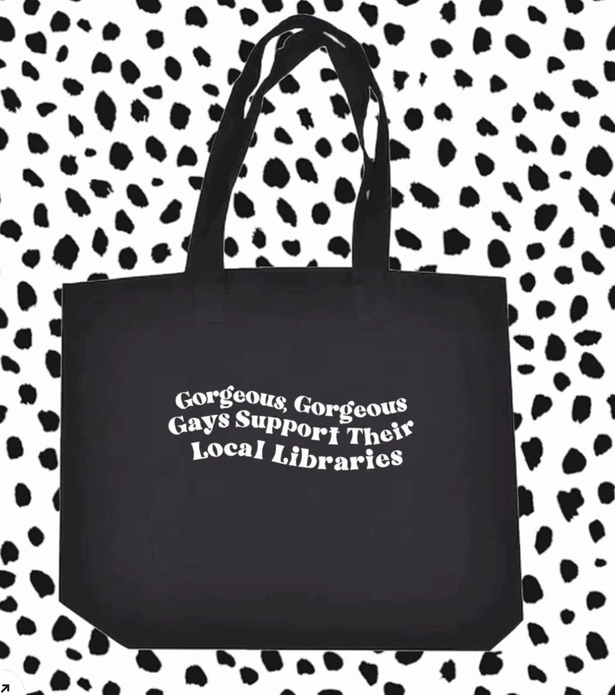 Image of a black tote bag on a black and white background. The tote bag has white lettering which reads "gorgeous gorgeous gays support their local libraries." 