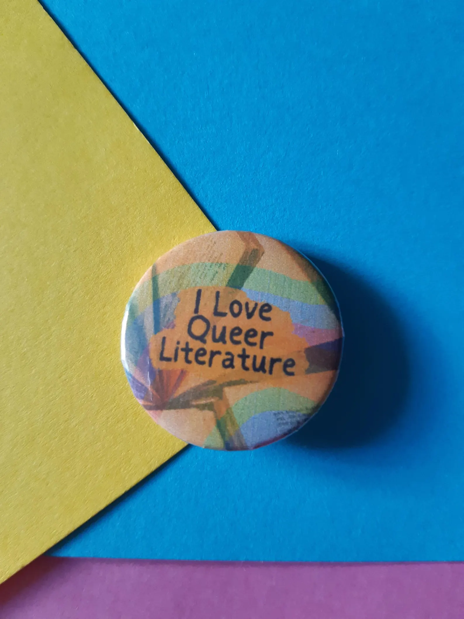 Image of a colorful button on a colorful background. The button reads "I love queer literature." 