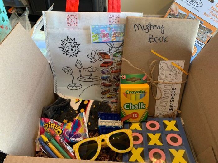 Picture of a cardboard box with sunglasses, crayons, a tote bag, a tic-tac-toe game, and a book wrapped in butcher block paper that says "Mystery Book," among other goodies 