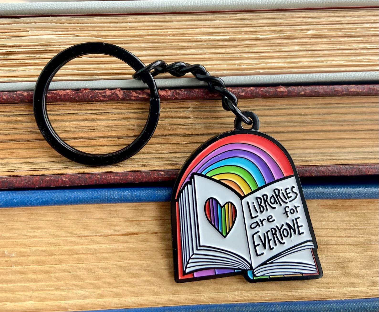 Image of a keychain on a stack of books. It features an open book with a rainbow and says "libraries are for everyone."