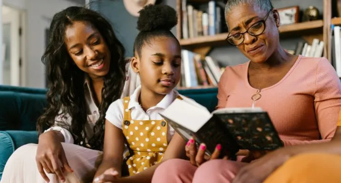 three generations of Black women (and one girl) reading a book on the couch
