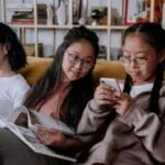 young Asian girls and one woman sitting together reading and talking on a couch
