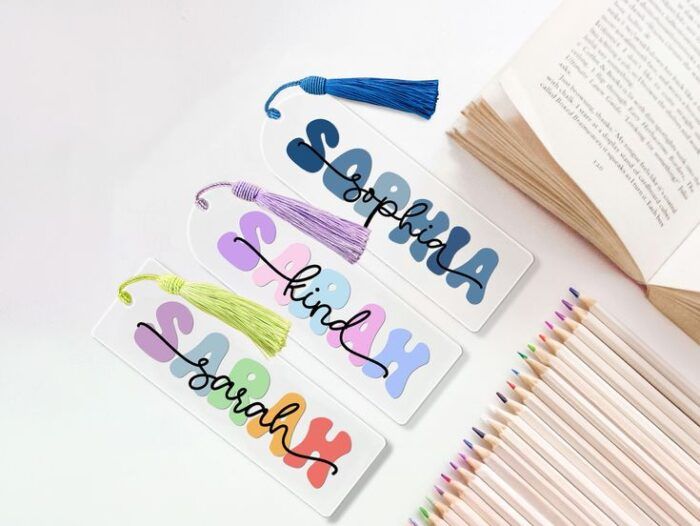 three bookmarks with different colored tassles. The design features a name in colorful bubble letters, layered under the same name in black script.