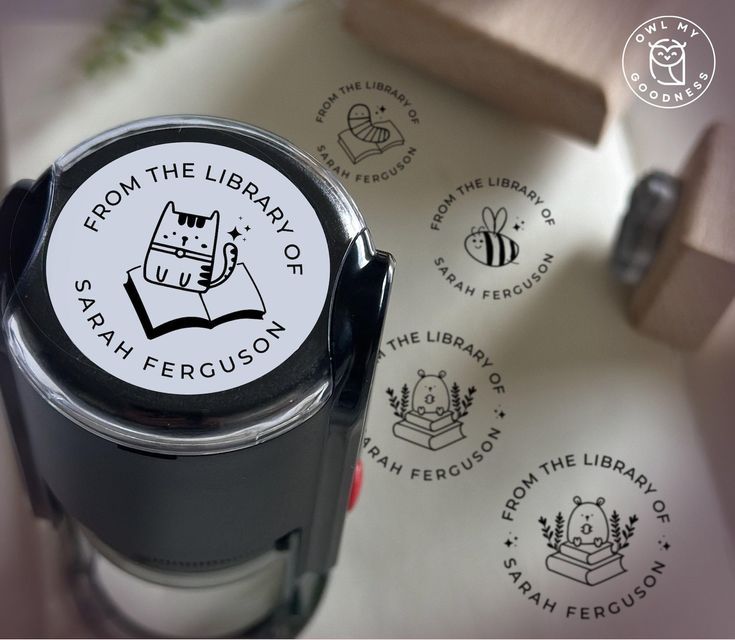 a circular self-inking stamp with the image of a cartoon cat reading and words around the edges reading "From the Library Of Sarah Ferguson" A piece of the paper in the background shows sample stamps with different cartoon images in the middle