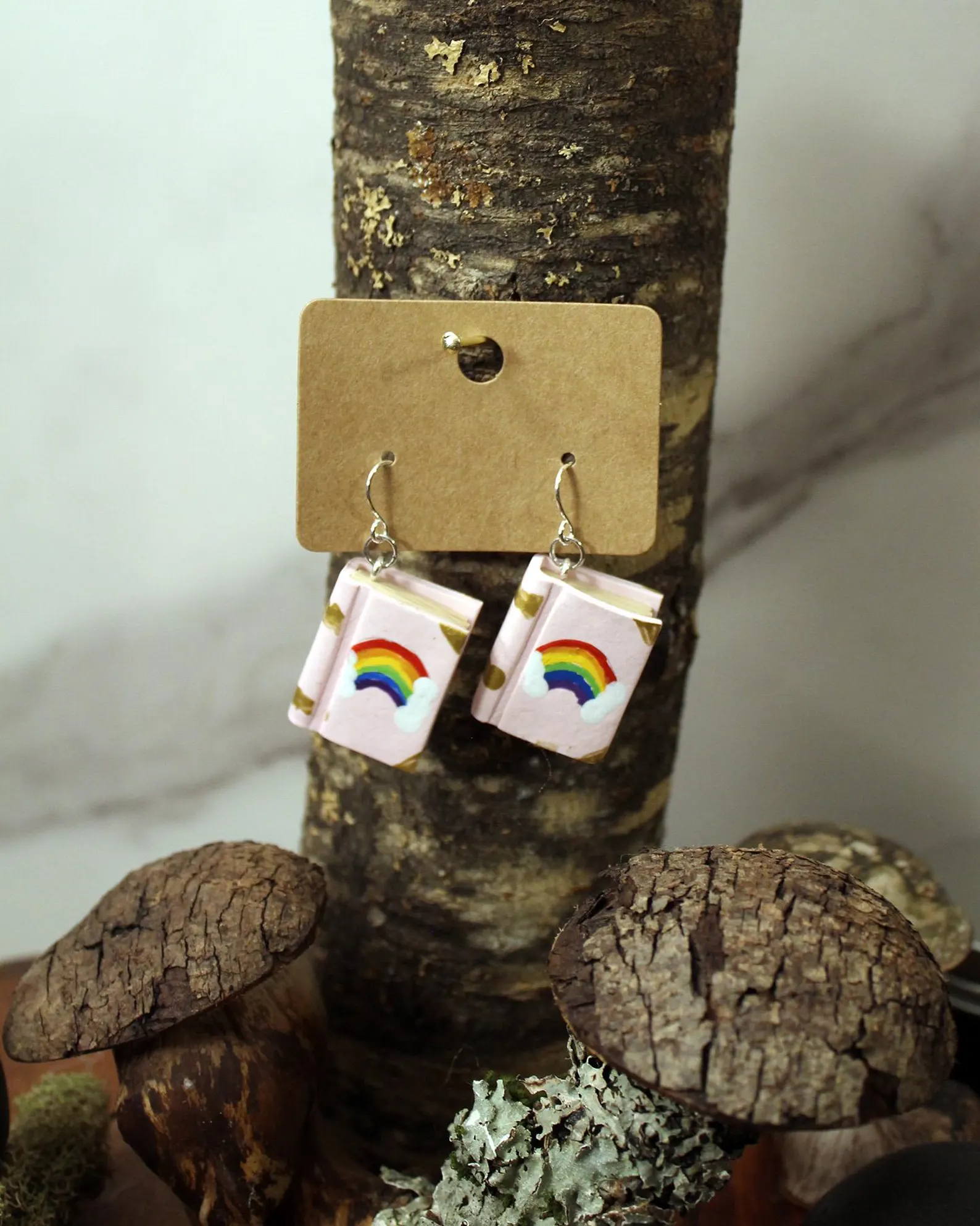 Image of a pair of polymer clay earrings. They are in the shape of hardcover books. The background is white with a rainbow at the center. 