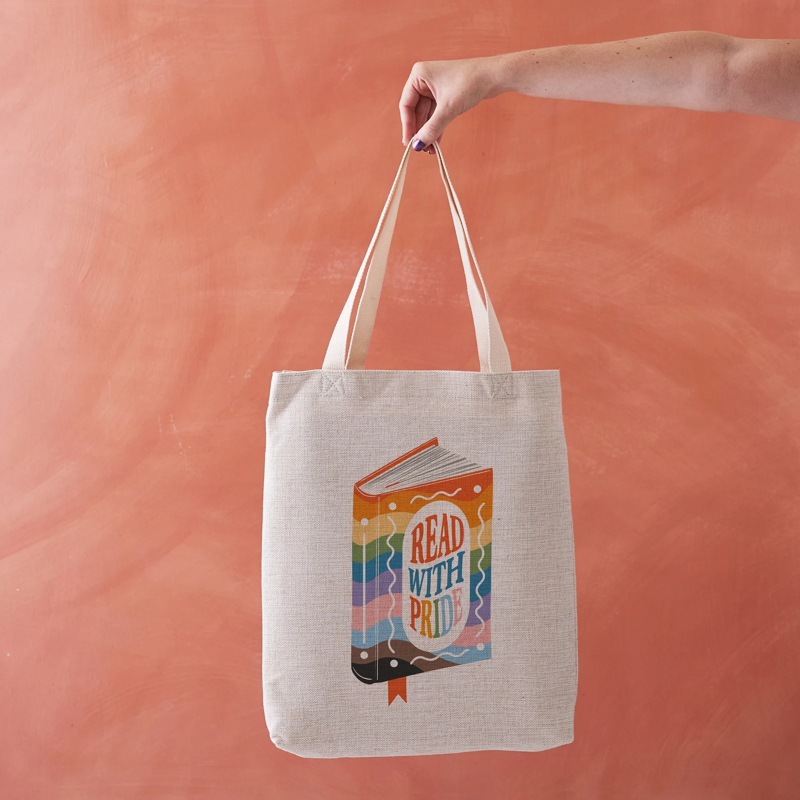 Image of a white hand holding a canvas tote bag against a peach colored background. The tote bag has a rainbow colored book that says "read with pride" on the front. 